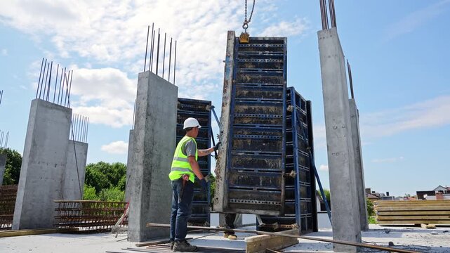 Two workers align tall metal formwork for a concrete wall in direct daylight, Hardhat laborers steady the panel on an elevated building slab