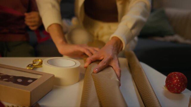 Close-up shot of unknown woman wrapping Christmas present with craft paper at table near child. Clip suitable for holiday marketing campaigns, DIY craft tutorials, and festive seasonal advertising