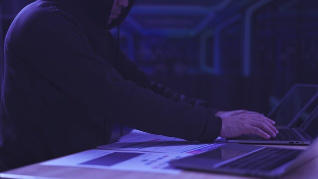 Hooded hacker types on laptop keyboard over data reports in dark room. Cybersecurity breach attack concept, corporate espionage identity theft threat, malicious software digital surveillance.