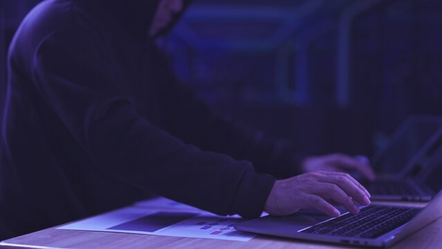 Hooded hacker types on computer keyboard in dark room near charts. Cybercrime security threat, network espionage attack, database malware breach defense, information technology system audit.