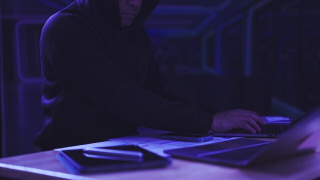 Hooded hacker types on laptop keyboard in dark server room. Cybersecurity threat defense, network espionage crime prevention, software ransomware extortion attack, data breach security monitoring.