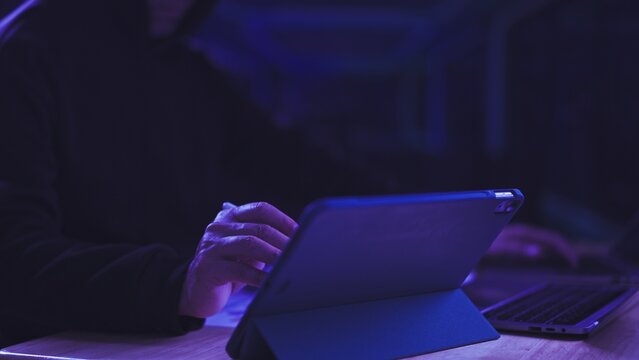 Hacker types on digital tablet computer in dark room. Cyber security threat concept, data breach surveillance, network system unauthorized access, malicious software espionage.