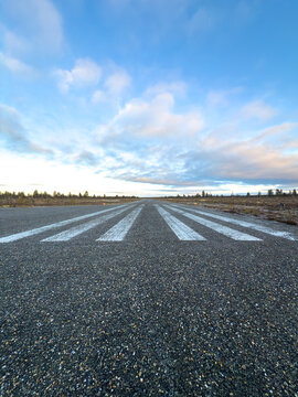 A low ground-level vertical shot looking along white painted threshold markings on an empty asphalt airfield runway in rural Finland under a bright sky.