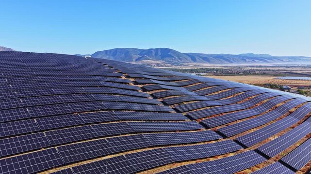 Solar panels cover the surface of a hillside in a rural location. Mountains rise in the background. The weather is sunny with no clouds in the sky.