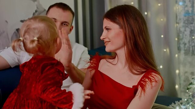 Young child plays with parents on a couch. The child wears a red dress, smiling at parents. Parents smile, looking at child with affection. Warm holiday lights frame the cozy scene