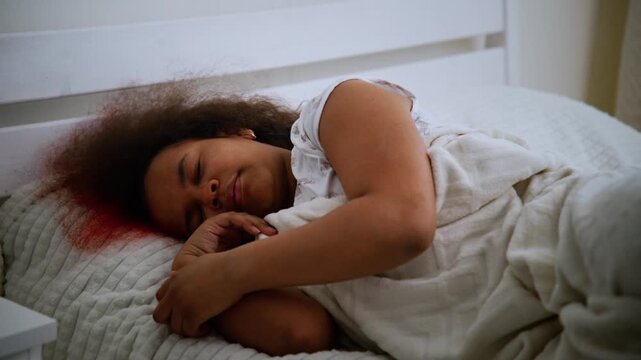 Girl sleeps peacefully in bed. Hair curls outward in soft sleep. Red streaks on pillow suggest recent injury. White bedding frames quiet, restful scene