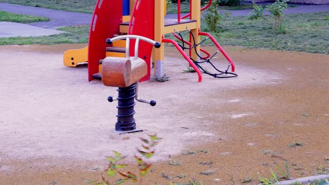 Wooden-and-metal spring rocker with a log-style seat and curved handrails stands on a beige sand surface; to the right, a colorful play structure features red panels, yellow supports, and climbing ele