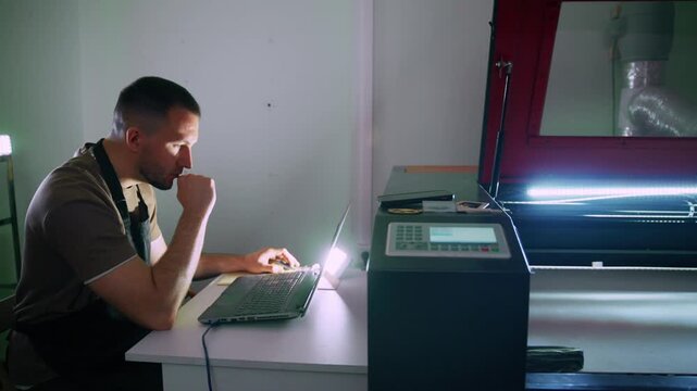 Man focuses intently on laptop in workshop. He wears apron, typing with concentration. Large machine glows beside him, active and ready. He checks files or designs, perhaps preparing for printing