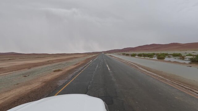 White car drives along a paved road in the Namib Desert; there is a lot of water around following the rain. Namibia