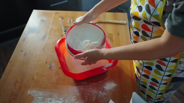 Hands sift flour into a red bowl. Wooden table holds scattered flour. Apron with colorful patterns adds charm. Sifting action suggests baking preparation. Ideal for cooking or home baking scenes