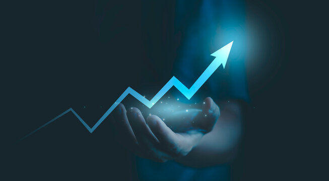 Success, progress, financial goals, and strategic development. Business growth concept showing a glowing upward arrow and rising line chart above an outstretched hand on dark blue background.