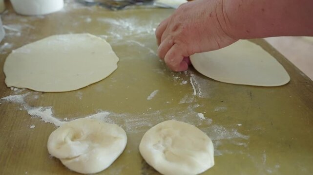 Hand shapes dough on floured wooden surface. Roller rests nearby, ready for flattening. Dough balls sit beside flattened circles. Warm kitchen setting, flour dusted everywhere. Ideal for food, cooking