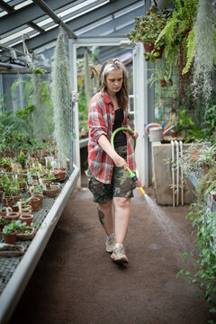 Hardworking woman botanist working in greenhouse watering pavilion ground and flowers with hose, focused female plant nursery employee taking care of plants and environment, spraying water over ground