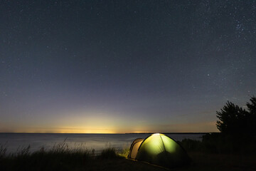 Illuminated camping tent under starry night sky on Baltic Sea coast in Estonia with distant city light pollution across horizon © Dmitri