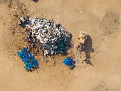 Aerial view of a scrap metal recycling yard with a large pile of assorted metal waste, a yellow front-end loader, and blue containers in Brighton, Colorado, United States.