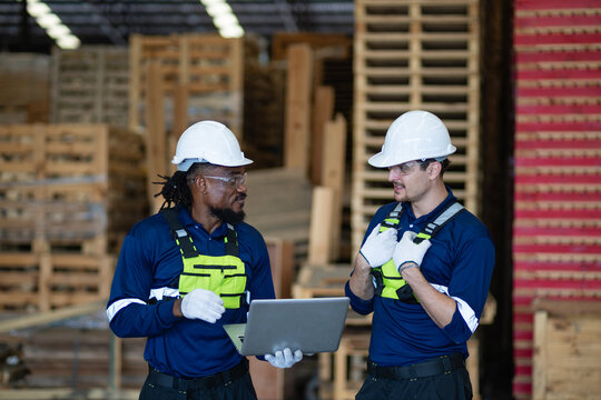 During teamwork discussions, two factory workers hold a team meeting while one man carries a laptop beside wooden pallets.