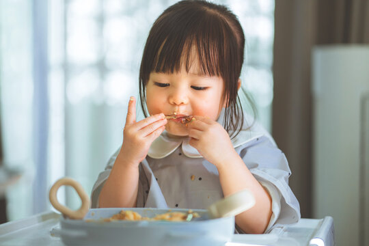 Toddler girl eating food with fingers while sitting in high chair at home, learning to feed herself independently during everyday mealtime, developing hand coordination.