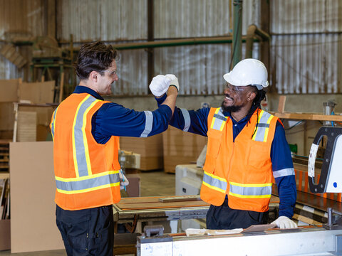 Pausing plywood woodworking, a carpenter and his colleague cross forearms in a friendly celebration beside a panel saw in the workshop.