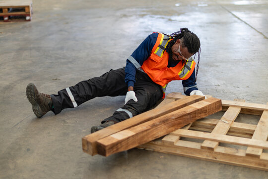 Man with workplace accident injury sits and clutches his leg in pain beside fallen wooden planks and a wooden pallet.