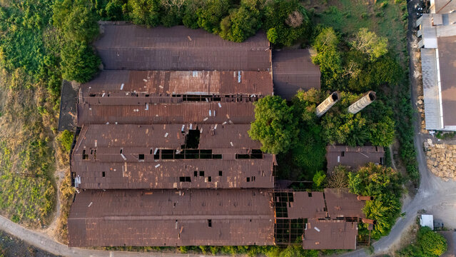 Aerial view of an abandoned industrial factory with a weathered, rusty corrugated metal roof, overgrown with lush green trees and vegetation, featuring two tall chimneys in Indonesia.