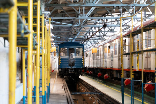 Blue vintage railcar viewed from behind between modern passenger trains inside a railway maintenance hall with yellow safety posts, metal beams and tracks.