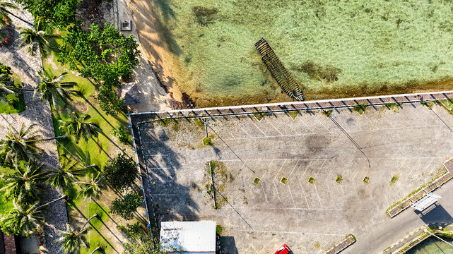 Aerial view of the Karimunjawa Passenger Ferry Port with a paved parking area, palm trees, and turquoise water with a small boat near the shore in Karimunjawa, Central Java, Indonesia.