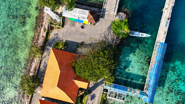 Aerial view of the Karimunjawa passenger ferry port with a long pier, turquoise water, and a building with a red roof in Karimunjawa, Central Java, Indonesia.