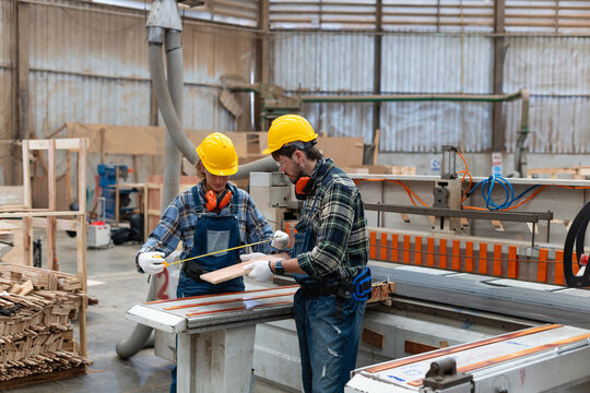 A woman practices carpentry in a workshop, measuring wood with a tape measure alongside her male colleague at a panel saw.