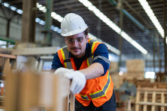 Inside a woodworking workshop, a carpenter pushes plywood pieces across a stack while looking toward the camera near wooden pallets.