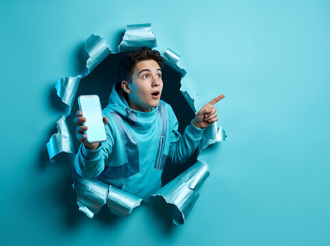 Surprised young man holding smartphone and pointing through torn blue paper