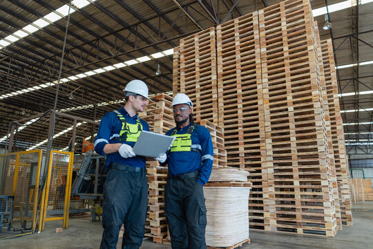 Wooden pallet stacks fill a warehouse logistics area where two colleagues look together at a laptop beside the pallets.