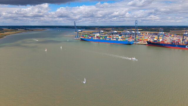 Aerial view of the Port of Felixstowe with large container ships docked at the terminal, towering cranes, and sailboats navigating the water in Felixstowe, England, United Kingdom.