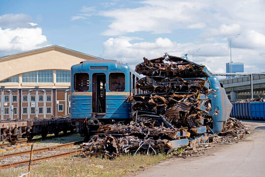 Old blue passenger carriages stand beside piles of dismantled railway materials in a rail yard, with exposed metal frames and industrial equipment, with visible signs of wear...