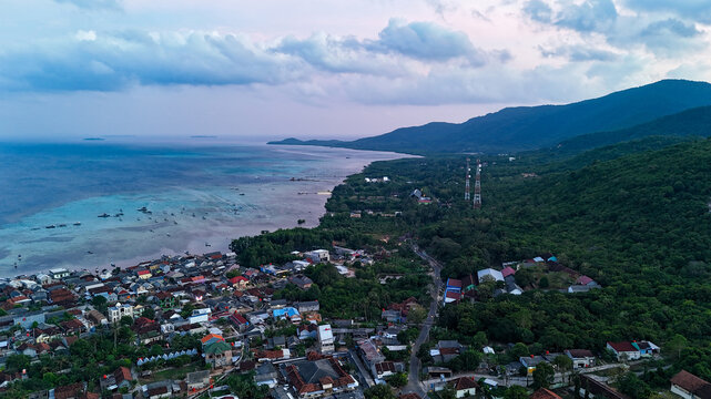 Aerial view of Karimunjawa showing the coastal village with dense houses, lush green hills, and turquoise waters of the Java Sea in Karimunjawa, Central Java, Indonesia.