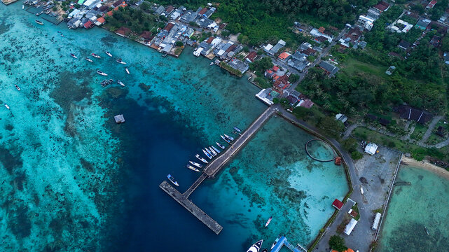 Aerial view of Port Karimunjawa with boats docked at the pier, surrounded by turquoise waters, coral reefs, and coastal buildings in Karimunjawa, Central Java, Indonesia.