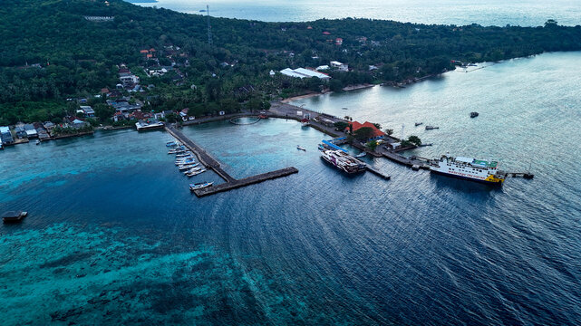 Aerial view of Port Karimunjawa with ferry boats docked at the pier, surrounded by turquoise tropical waters, lush green hills, and coastal buildings in Karimunjawa, Central Java, Indonesia.