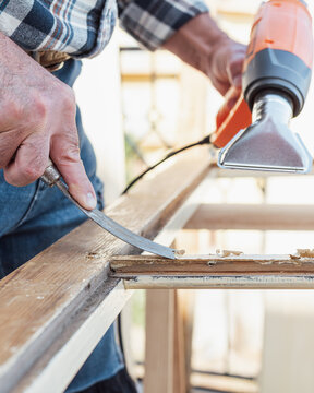 Carpenter using heat gun and wood scraper removes paint from old wooden window. Construction industry, carpentry. Restoration. 