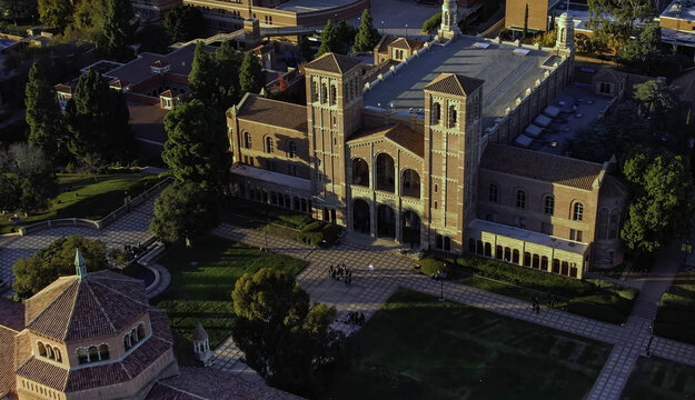 Aerial view of the historic Royce Hall on the UCLA campus, featuring its iconic twin towers, red-tiled roof, and the surrounding paved courtyard with students, Westwood, California, United States.