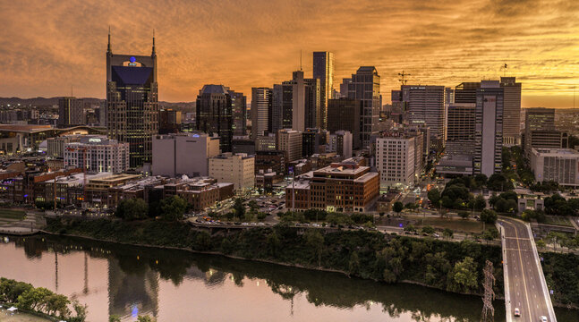 Aerial view of the downtown Nashville skyline at sunset, featuring the Cumberland River, modern skyscrapers, and vibrant orange sky reflecting on the water in Nashville, TN, United States.