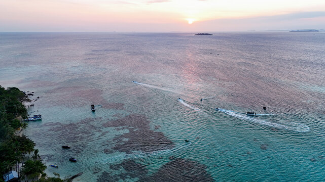Aerial view of Pantai Tanjung Gelam at sunset with boats leaving wakes in the turquoise water over coral reefs and lush trees along the shoreline in Karimunjawa, Central Java, Indonesia.