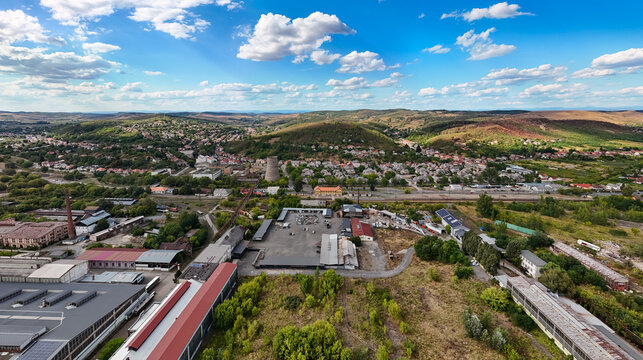 Aerial view of the former metallurgical works and industrial buildings beside railway tracks in Ozd, Hungary, surrounded by homes, wooded hills and rolling countryside.