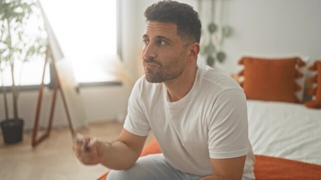 Man holding creditcard to chin while sitting on bed in bedroom next to window and plant; quiet contemplation.