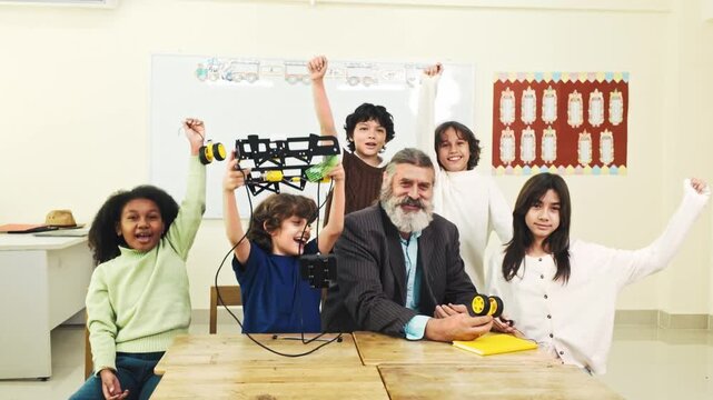 Smiling students and a teacher celebrate a classroom robotics project, holding wheels and a frame at a school desk