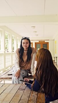 Two women sit at a wooden table in a bright corridor; one looks upset while the other listens, offering support during an emotional conversation.