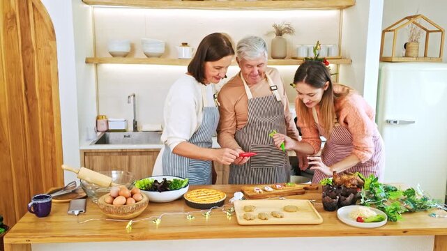Three people in a bright kitchen decorate holiday cookies together, wearing aprons amid bowls, eggs, and greenery for a festive homemade dessert spread.