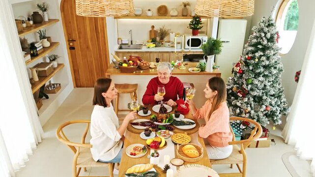 Three people share a festive Christmas meal in a cozy modern kitchen, toasting at a decorated table beside a lit tree and seasonal treats.