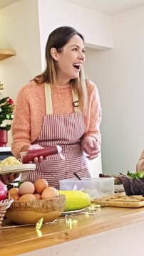 Smiling woman in a striped apron cooks in a cozy home kitchen, holding a sauce bottle beside eggs, fruit, and ingredients on a wooden counter during a festive gathering.