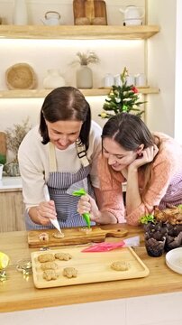 Two women decorate gingerbread cookies with icing in a cozy kitchen, with holiday decor and a small Christmas tree in the background
