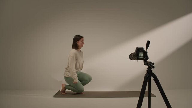 Woman moving through yoga stretch poses beside camera tripod in minimal studio during production set