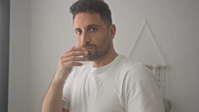 Man smiling with hand to cheek in studio apartment, white tshirt, beard and gentle pose near macrame wall hanging; calm confidence.
